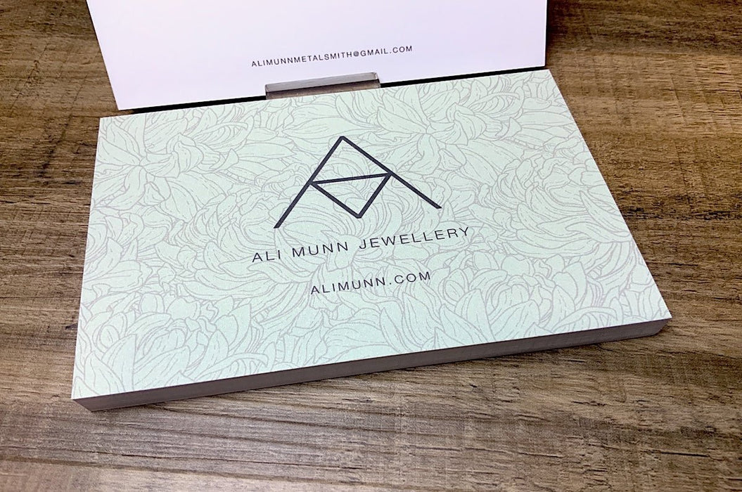 Custom 14pt uncoated matte postcards with light blue floral design and Ali Munn Jewellery logo on a wood grain background