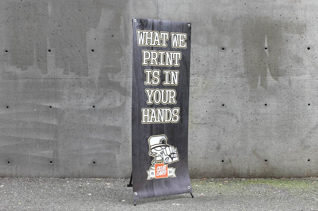 Angled view of a 24x60 outdoor X banner stand with custom printed black graphic set up against a concrete wall.  Clubcard Printing USA