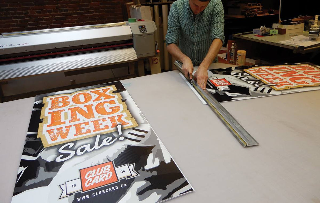Print shop technician trimming a full-color printed vinyl replacement banner for an X-stand or L-stand display. Clubcard Printing USA