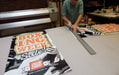 Print shop technician trimming a full-color printed vinyl replacement banner for an X-stand or L-stand display. Clubcard Printing USA