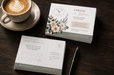 A styled flat-lay of printed coffee shop marketing cards with gold foil stamped text and a floral product box on a dark wooden table, displayed alongside a cup of latte art and a pen, printed by Clubcard Palm Springs on 12pt 100% recycled fibers stock with full color detail.